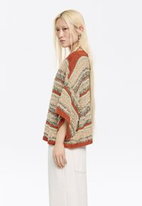 Gestrickter, mehrfarbiger Pullover mit kurzen Ärmeln, der wellenförmige Streifen in Rost-, Grün- und Cremetönen zeigt. Das Model trägt einen hellen Rock.