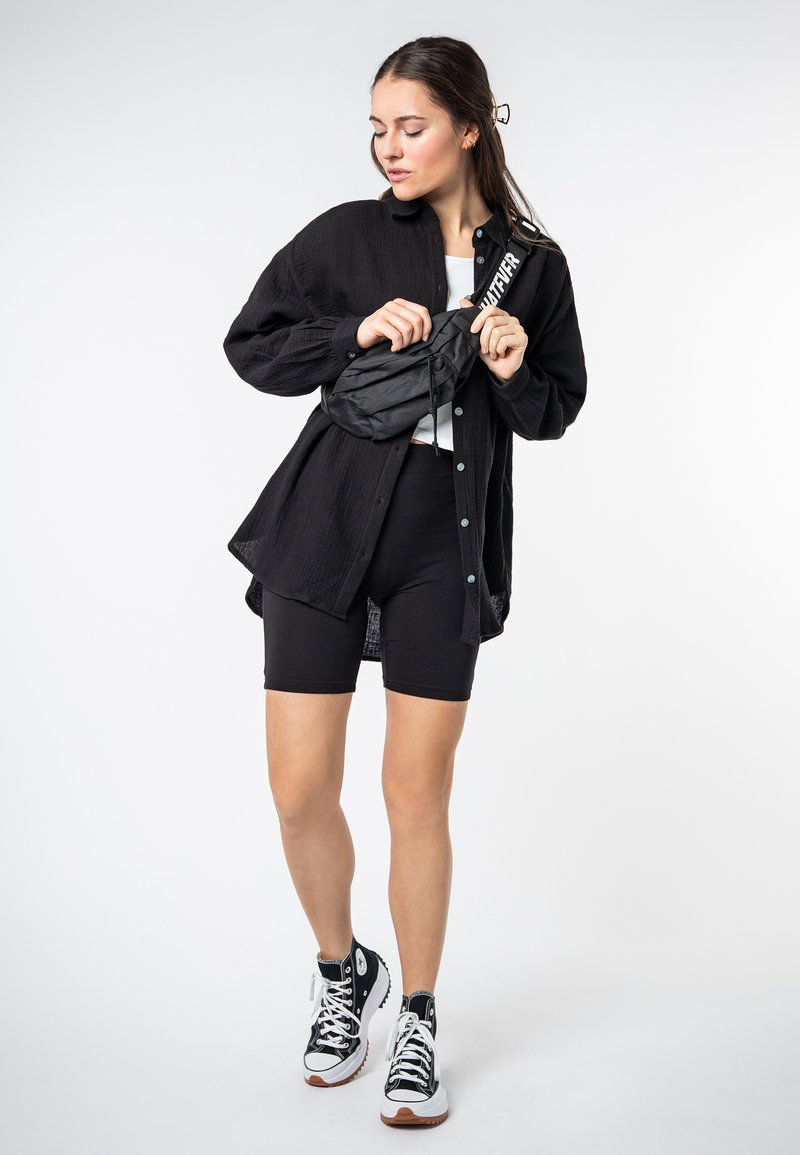 Oversized black button-up shirt, white cropped top, black biker shorts, black fanny pack with white strap, and black high-top sneakers.