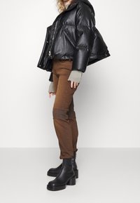 Chaqueta de plumas negra con acabado brillante y capucha, combinada con pantalones ajustados marrones con parches en las rodillas y botas de plataforma negras hasta el tobillo.