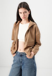 Veste à capuche zippée marron avec une finition texturée, dotée de poches avant et de poignets ajustés, assortie à un jean large bleu clair.