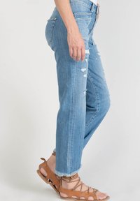 Jeans en denim bleu clair avec des détails usés, une coupe droite et des ourlets effilochés, associés à des sandales à lanières beiges.