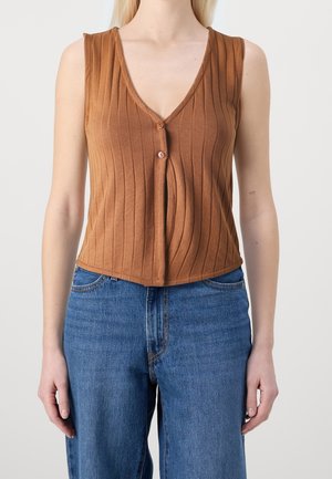 Mujer vistiendo una blusa sin mangas de punto marrón con botones, combinada con unos vaqueros de tiro alto azul, de pie frente a un fondo liso.