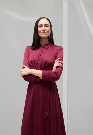 Μπορντό φόρεμα με κουμπωτό γιακά, μακριά μανίκια και πλισέ σχέδιο. Ομαλή υφή, που φτάνει μέχρι το γόνατο, με λεπτομέρειες δεμένης μέσης.