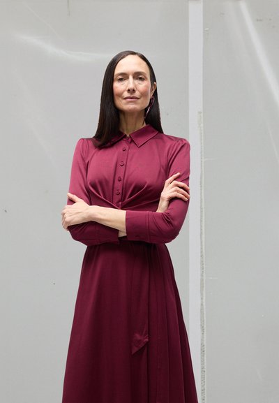 Bordeauxrotes Kleid mit einem geknöpften Kragen, langen Ärmeln und einem plissierten Design. Glatte Textur, knielang, mit einem Taillendetail zum Binden.