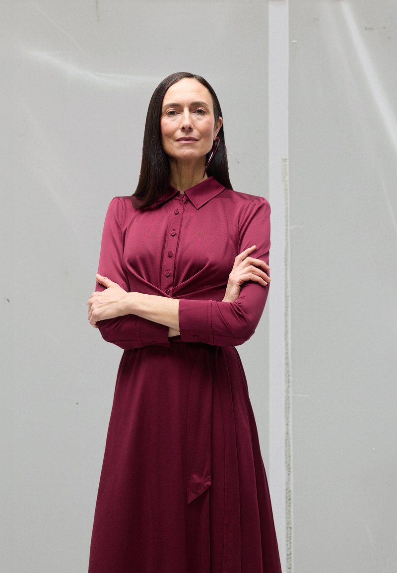 Bordo haljina s ovratnikom na kopčanje, dugim rukavima i naboranim dizajnom. Glatka tekstura, seže do koljena, s detaljem vezanja u struku.