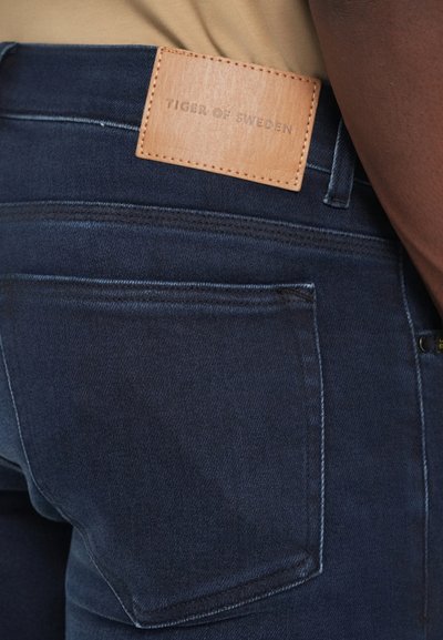 Jeanși din denim de culoare albastru închis, cu o textură netedă, având un eticheta de brand din piele bej cusută pe talie și un design clasic cu cinci buzunare.