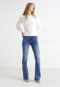 Blusa blanca texturizada con mangas abullonadas, combinada con jeans de mezclilla azul ajustados y acampanados. La modelo está de pie contra un fondo sencillo.