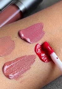 Neljä huulikiillon näytettä vaaleanpunaisissa ja punaisissa sävyissä iholla, valkoinen aplikaattori levittää kirkkaan punaista kiiltoa.