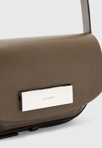 Ruskea nahkainen olkalaukku, jossa on suorakulmainen muoto, sileä pinta ja hopeinen metallinen yksityiskohta, jossa on "ALLSAINTS" kaiverrus.