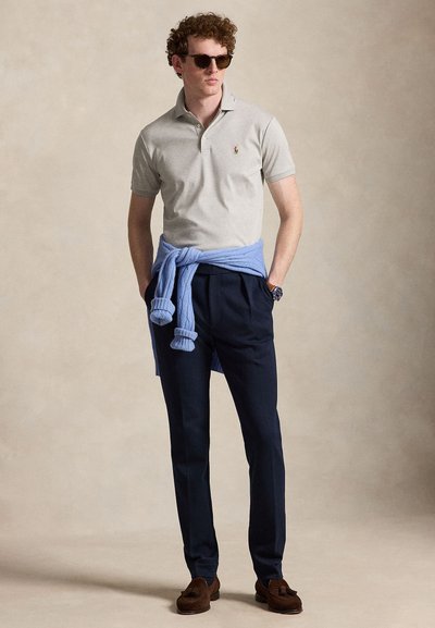 Homme portant un polo gris, un pantalon bleu marine, un pull bleu noué autour de la taille, des mocassins marron, des lunettes de soleil et une montre-bracelet, debout les mains dans les poches.