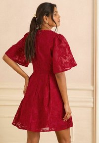 Piros csipke ruha rövid, bővülő ujjal, testéhez simuló fazonnal és A-vonalú szoknyával. Az anyag virágmintával és áttetsző textúrával rendelkezik.