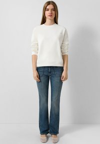 Biała bluza z mankietami, w połączeniu z niebieskimi dżinsami w kształcie dzwonów. Model nosi jasne buty. Prosty design i luźny krój.