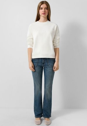 Weißes Sweatshirt mit aufgeschlagenen Ärmeln, kombiniert mit blauen Bootcut-Jeans. Modell trägt hellfarbene Schuhe. Schlichte Gestaltung und entspannter Sitz.