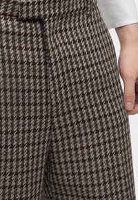 Gros plan sur un pantalon à motif pied-de-poule marron et noir avec un bouton et une partie d'une main reposant à côté de la hanche.