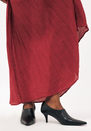 Persona con una falda larga roja texturizada y zapatos negros de tacón con punta afilada, de pie sobre un fondo blanco.