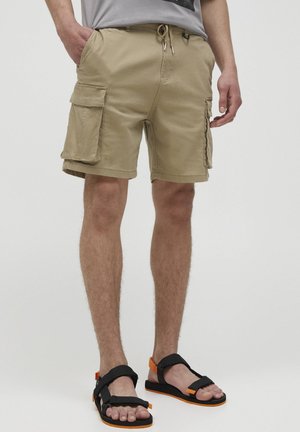 Homme portant un short cargo beige avec de grandes poches latérales et des sandales noires avec des accents orange, debout devant un fond uni.