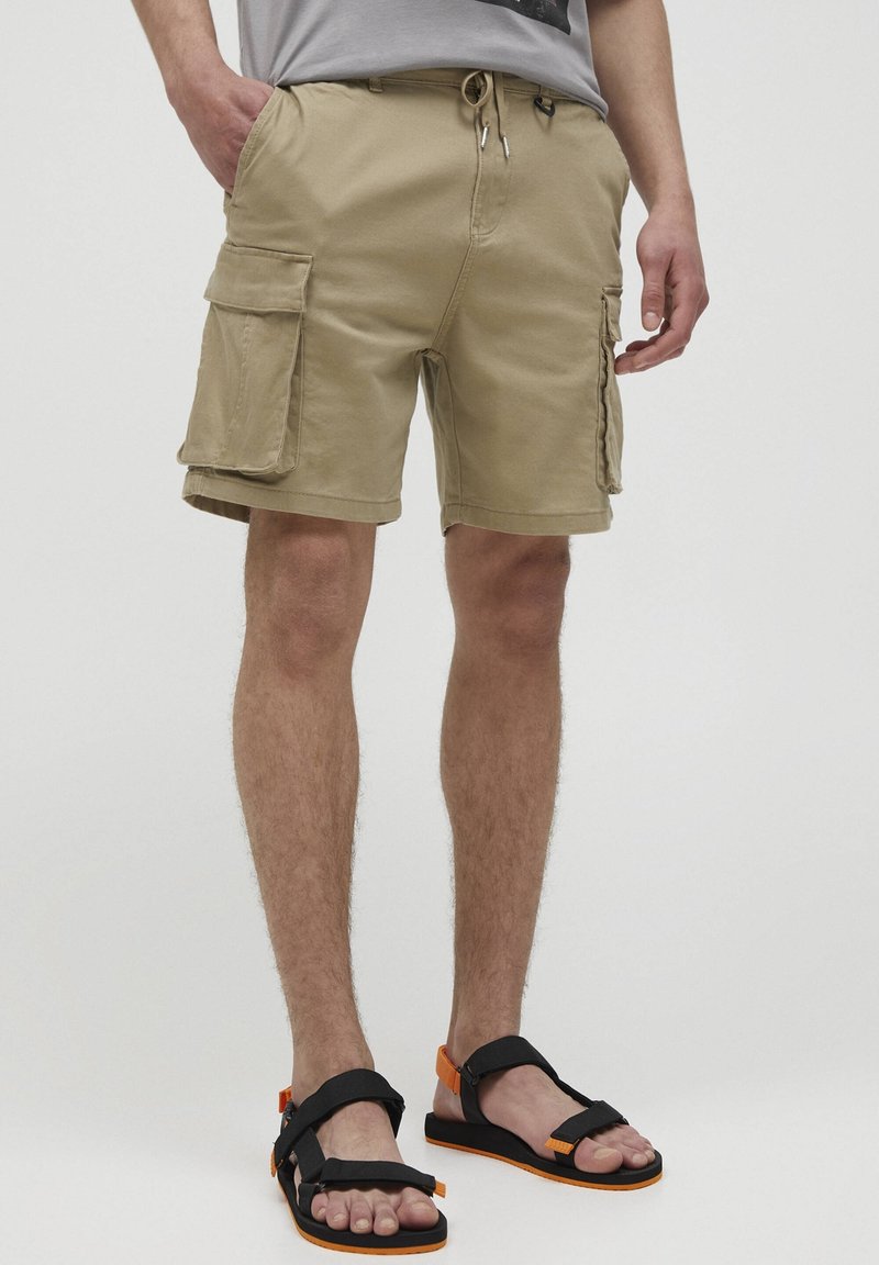 Homme portant un short cargo beige avec de grandes poches latérales et des sandales noires avec des accents orange, debout devant un fond uni.