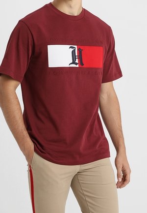T-shirt en coton rouge à manches courtes, comportant un panneau graphique blanc et rouge avec des lettres noires. Porté avec un pantalon beige avec des accents rouges.
