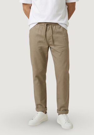 Uomo che indossa pantaloni beige con coulisse e risvolti arrotolati, e sneaker bianche, in piedi contro uno sfondo chiaro e uniforme.