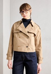 Heledbeez värvi lühendatud trench coat, millel on topelt nööpide rida, suured kraed ja metallist nööbid. Kantud musta kõrge kaelusega särke peal.