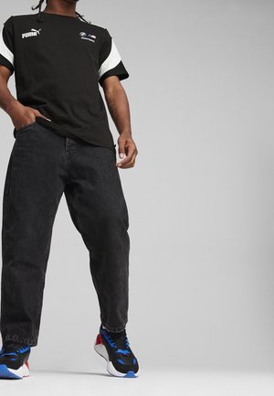 T-shirt noire avec des accents blancs, arborant les logos Puma et BMW. Associée à un jean noir ample et des baskets noires avec des accents bleus et rouges.