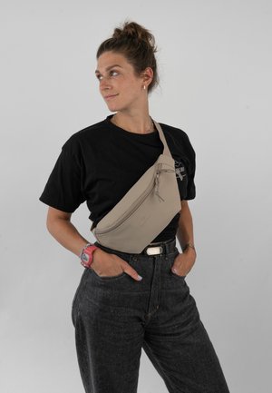 Borsa a tracolla beige in materiale morbido, con chiusura a zip, tracolla regolabile e branding discreto. Indossata sopra una maglia nera.