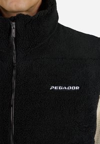 Črna flis brezrokavnica z visokim ovratnikom, sprednjim zadrgo in belim logotipom "PEGADOR" na prsih; prijeten na dotik in prilegajoč se.