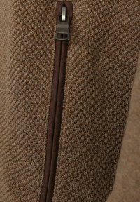 Strukturiertes braunes Gewebe mit einem Strickmuster, das einen dunkelbraunen Reißverschluss auf einer Seite aufweist. Das Material wirkt warm und langlebig.