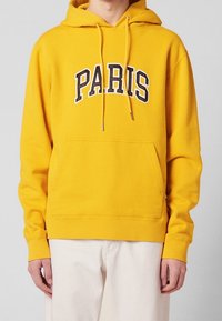 Sweat à capuche jaune avec l'inscription "PARIS" en lettres noires et blanches audacieuses. Il possède une poche avant et des poignets côtelés. Texture lisse.