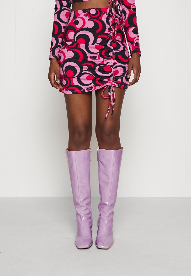 Femme portant une mini-jupe à motifs roses, noirs et rouges avec des cordons latéraux et des bottes brillantes lavande montant jusqu'aux genoux.
