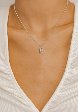 Silberne Halskette mit einem Anhänger in Form eines großen "M" mit strukturiertem Finish, der auf nackter Haut über einem weißen, gerippten Oberteil liegt.