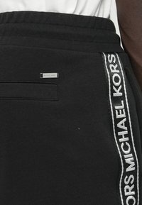 Czarne spodnie dresowe wykonane z miękkiego materiału, z ściągaczem w pasie, boczną kieszenią oraz białymi paskami z logo po bokach.