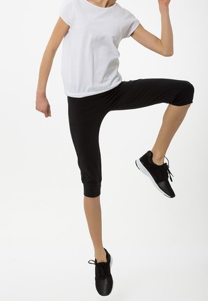 Osoba ubrana w czarne krótkie legginsy i czarne trampki, unosząca jedno kolano wysoko, ubrana w biały top z krótkim rękawem.