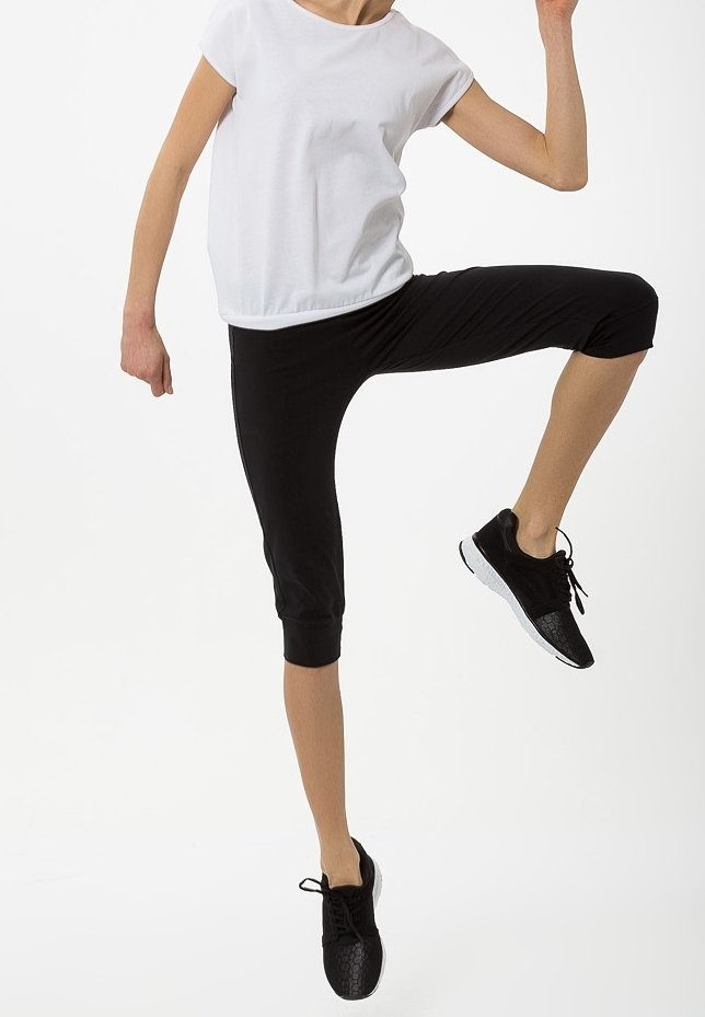 Person iført sorte korte leggings og sorte sneakers, løfter det ene knæ højt, iført en hvid top med korte ærmer.