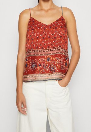 Rdzawo-czerwona kwiecista koszulka na ramiączkach z cienkimi paskami, noszona z kremowymi, wysokim stanem spodniami, jedna ręka w kieszeni na jednolitym tle.