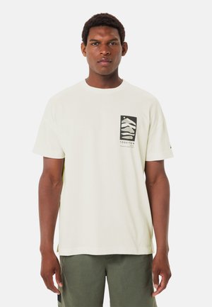 Uomo con una T-shirt bianca con una grafica rettangolare nera e testo sul petto, abbinata a pantaloni verde oliva, in piedi su sfondo bianco.