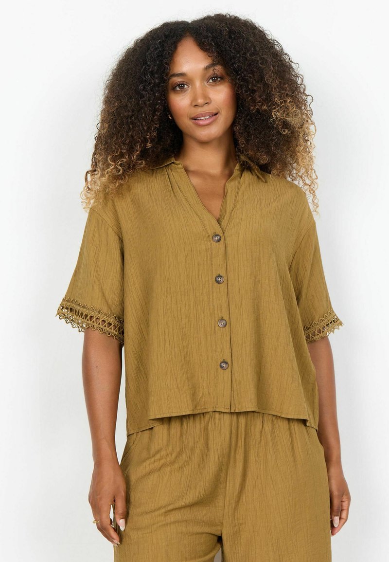 Femme aux cheveux bouclés portant une chemise boutonnée ample couleur moutarde avec des manches courtes bordées de dentelle et un pantalon assorti, sur fond blanc.