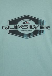 Světle tyrkysové tričko vyrobené z bavlny, s výrazným černým hexagonálním logem Quiksilver a bílým textem a grafiku vlny.