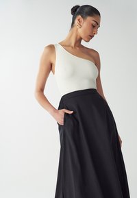 Top bianco a costine con una spalla, abbinato a una lunga gonna nera con tasche laterali, texture liscia e silhouette classica.