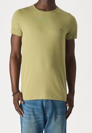 Homme portant un t-shirt vert olive clair à manches courtes avec un petit logo sur la poitrine et un jean en denim bleu, debout devant un fond blanc.