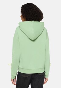 Miętowa bluza z kapturem, z sznurkiem do regulacji, ściągaczami przy mankietach i bocznymi wiązaniami na rękawach, wykonana z miękkiego materiału. Charakteryzuje się luźnym krojem.