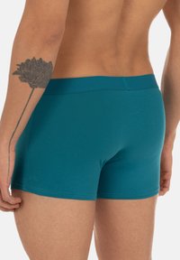 Boxer in cotone color turchese con vita senza cuciture, design aderente e texture liscia. Tatuaggio visibile sul braccio sinistro.