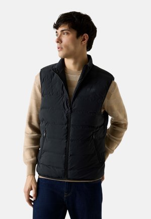 Giovane uomo che indossa un maglione beige a maglia e un gilet imbottito nero con cerniera, che guarda a sinistra su uno sfondo neutro.