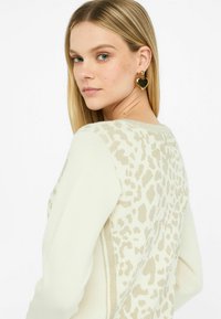 Maglione chiaro con stampa leopardata beige sulla schiena, con scollatura rotonda e maniche lunghe. Sono indossati orecchini a forma di cuore dorati.