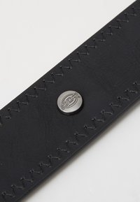 Correia de pele preta com costura visível e um botão redondo prateado com o logótipo da Dickies. Superfície suave com acabamento mate.