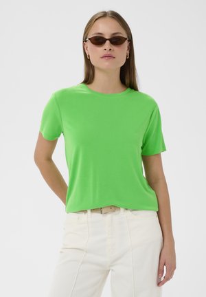 Žena sohnutá v hnedom slnečnom okuliaroch, zelenom tričku s krátkym rukávom, bielych nohaviciach s béžovým opaskom, stojaca pred jednofarebným svetlým pozadím.