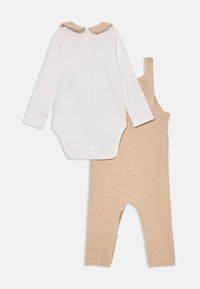 Bijeli baby bodysuit dugih rukava s bež ovratnikom i bež pletivom hlačama s naramenicama raširenim na bijeloj podlozi.