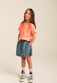 Lysende neon pink bomulds T-shirt, korte ærmer, med et lille broderet design; parret med en lyseblå denim nederdel og sneakers.