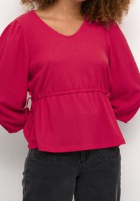 Fuchsia tekstureret top med V-hals, samlede talje og bløde ærmer. Sammen med mørke jeans for et afslappet look.