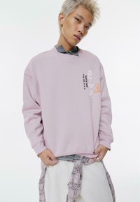 Felpa rosa chiaro realizzata in tessuto morbido, con collo a giro, spalle scese e grafiche stampate in colori a contrasto.